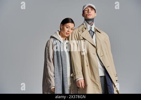 Un homme et une femme élégants se tiennent gracieusement côte à côte dans des trench-coat sur un fond de studio gris. Banque D'Images