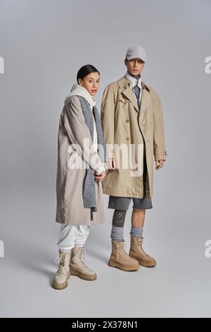 Jeune couple élégant en trench-coat debout côte à côte dans un studio, exsudant l'élégance urbaine sur un fond gris. Banque D'Images