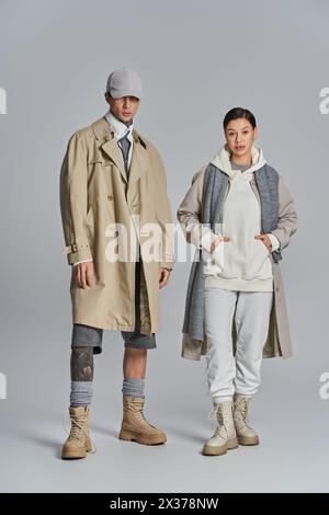 Un jeune couple élégant en trench-coat debout à côté l'un de l'autre dans un studio, sur un fond gris. Banque D'Images
