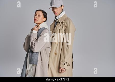 Un jeune couple élégant en trench-coat debout l'un à côté de l'autre dans un studio sur un fond gris. Banque D'Images