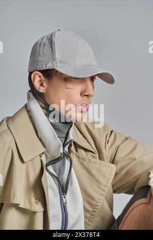 Un jeune homme tatoué se tient confiant dans un trench-coat et un chapeau, dégageant un air de mystère et d'intrigue sur un fond gris. Banque D'Images