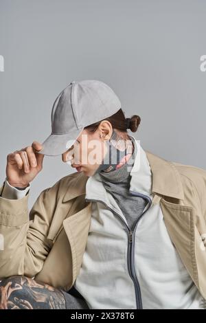 Un jeune homme avec un tatouage sur son visage est assis, vêtu d'un trench-coat, dans un studio sur un fond gris. Banque D'Images