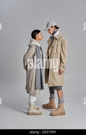 couple se tient côte à côte dans des trench-coat élégants, dégageant une ambiance sophistiquée et à la mode sur un fond gris. Banque D'Images
