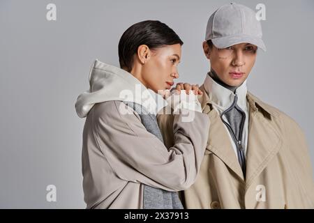 Un jeune couple élégant se tient gracieusement à côté de l'autre dans des trench-coat sur un fond de studio gris. Banque D'Images