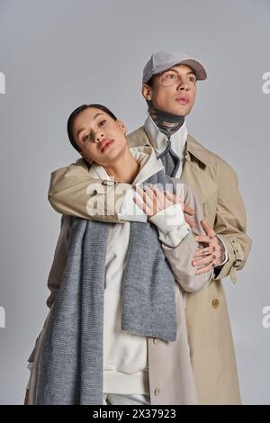 Un jeune homme et une jeune femme se tiennent gracieusement côte à côte dans des trench-coat, dégageant sophistication et style, sur fond de studio gris. Banque D'Images