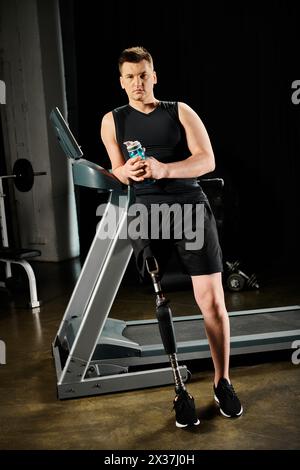 Un homme se tient debout sur un tapis roulant, tenant un verre dans sa main tout en s'entraînant au gymnase avec une jambe prothétique. Banque D'Images