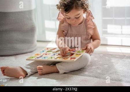 Image horizontale d'une petite fille aux cheveux bouclés jouant assise sur le tapis dans sa chambre avec un alphabet en bois, matériel Montessori. concept devel Banque D'Images