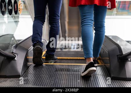 Jeune couple latino-américain méconnaissable, marchant ensemble dos à dos pour monter l'escalator. Tir court moyen de leurs jambes. Concept récréatif Banque D'Images