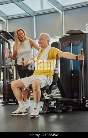 Femme senior active, avec la bataille de l'eau motivant l'homme faisant des exercices pour les mains, soulève des poids lui parle à la salle de gym moderne. Banque D'Images