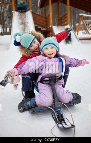 Rire petite fille et garçon assis sur le traîneau après la descente de toboggan en bois recouvert de neige le jour d'hiver. Banque D'Images