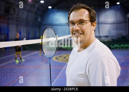 Homme souriant dans des lunettes avec raquette de badminton près du filet au terrain de sport. Banque D'Images