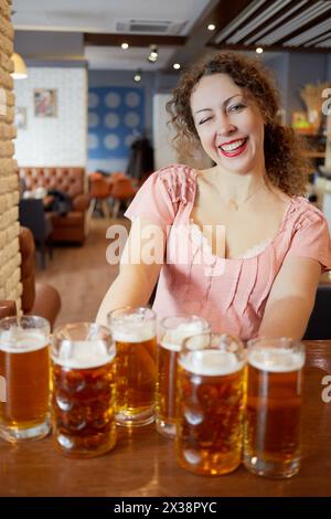 Portrait de femme souriante et clin d'œil avec les cheveux bouclés tient plusieurs tasses en verre avec de la bière dans les deux mains. Banque D'Images