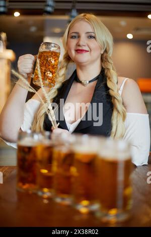 Femme blonde souriante se tient debout tenant des épillets dans une main et tasse de bière dans un autre au comptoir du bar dans le café, se concentrer sur la tasse. Banque D'Images