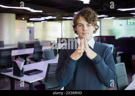 Beau adolescent pose avec les mains pliées dans un bureau moderne avec de nombreux lieux de travail Banque D'Images