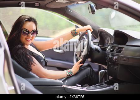 Femme heureuse avec de longs cheveux noirs dans des lunettes de soleil assis derrière le volant d'une voiture Banque D'Images