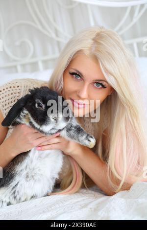 Jolie fille se trouve sur le lit blanc et tient le lapin noir moelleux dans la chambre Banque D'Images