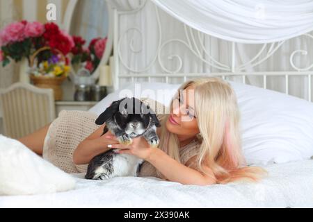 Jolie blonde se trouve sur le lit blanc et tient le lapin noir moelleux dans la chambre Banque D'Images