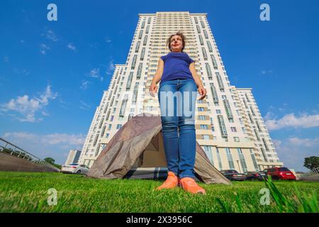 Belle femme se tient sur l'herbe près de tente, de grands bâtiments et des voitures à la journée ensoleillée Banque D'Images