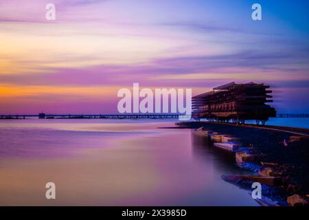 Image époustouflante capturant une structure architecturale moderne sur un coucher de soleil vibrant aux teintes violettes et roses reflétant un front de mer serein. Banque D'Images