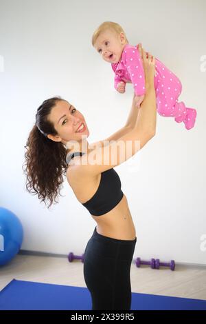 Mère dans les vêtements de sport debout ramasse bébé près dans la salle de gym Banque D'Images