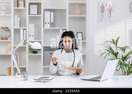 Une femme médecin hispanique analysant une lettre avec une expression inquiète dans son bureau, suggérant la réception de nouvelles inattendues ou négatives. Banque D'Images