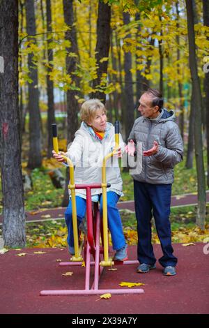 Les femmes âgées sont engagées sur le simulateur de sport et l'homme parle avec elle dans le parc d'automne avec des feuilles mortes Banque D'Images