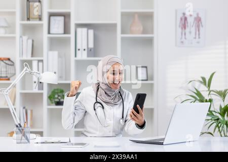 Heureuse médecin musulmane en hijab célébrant le succès tout en regardant son smartphone dans un bureau de clinique moderne. Banque D'Images