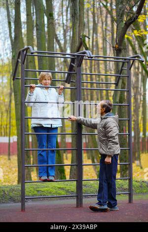 Les femmes âgées grimpent sur l'échelle et l'homme se tient près d'elle sur le terrain de jeu dans le parc d'automne Banque D'Images