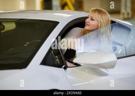 Jeune femme blonde regarde depuis le siège du conducteur de la voiture blanche moderne au parking souterrain. Banque D'Images