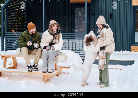 Parents caucasiens modernes assis sur une table en bois à l'extérieur tenant des tasses avec une boisson chaude regardant leur fille jouer avec le chien le jour d'hiver Banque D'Images