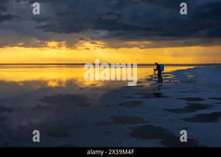 Photographe masculin se tient sur un lac tranquille pendant le coucher du soleil, capturant les paysages à couper le souffle où le ciel et l'eau se rencontrent, reflétant des couleurs vibrantes et des nuages spectaculaires. Banque D'Images