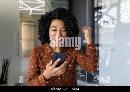 Une jeune femme joyeuse dans une élégante chemise à pois marron célèbre le succès tout en regardant son smartphone dans un espace de bureau moderne, exprimant le bonheur et la réussite. Banque D'Images