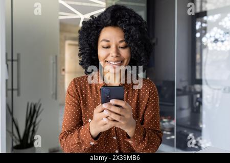 Une jeune femme joyeuse se tient dans un bureau élégant, engrossée dans son smartphone. Son engagement véhicule la connectivité et le professionnalisme dans un environnement d'entreprise détendu. Banque D'Images