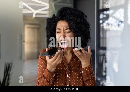 Femme afro-américaine dans une chemise à pois exprimant la colère et la frustration tout en criant dans un téléphone portable dans un cadre de bureau moderne. Banque D'Images