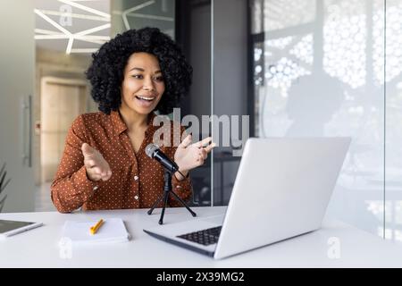 Une jeune femme afro-américaine joyeuse enregistrant un podcast dans un bureau moderne bien éclairé. Elle parle animée dans un micro, exprimant passion et professionnalisme. Banque D'Images