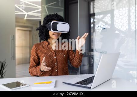 Une jeune femme aux cheveux bouclés s'engage avec la technologie de réalité virtuelle dans un espace de travail moderne. Elle fait des gestes en plein air tout en portant un casque VR, avec un ordinateur portable et un smartphone sur le bureau. Banque D'Images