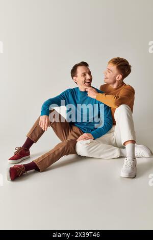 Deux hommes assis sur le sol, riant joyeusement en tenue décontractée sur fond gris. Banque D'Images