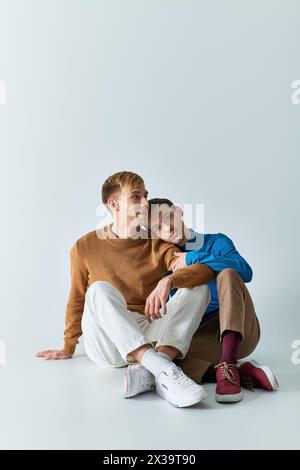 Deux hommes dans des vêtements décontractés assis sur le sol avec les bras l'un autour de l'autre sur fond gris. Banque D'Images