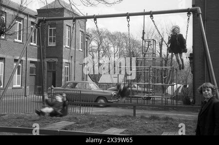 Années 1960, historique, trois jeunes écolières dehors dans une aire de jeux d'école, deux d'entre elles jouant sur une balançoire traditionnelle à la maille, Angleterre, Royaume-Uni. Une voiture de l'époque stationnée à côté d'un cadre d'escalade. Banque D'Images