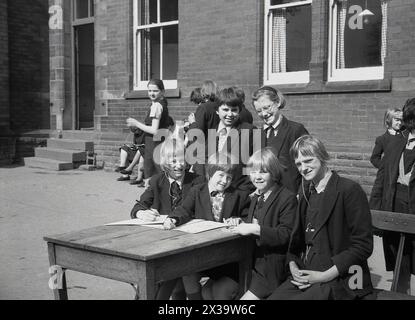 Les années 1960, des écolières historiques, du début de l'été et catholiques romains dans leurs uniformes assis à l'extérieur de leur bâtiment scolaire, avec un stylo et du papier, faisant leur travail scolaire sur un bureau en bois, Angleterre, Royaume-Uni. Banque D'Images