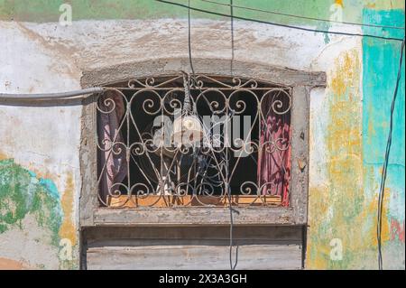 Grille de ferronnerie sur une fenêtre au-dessus d'une porte, entourée de peinture décolorée et pelée dans les rues arrière de la vieille ville, la Havane, Cuba. Banque D'Images