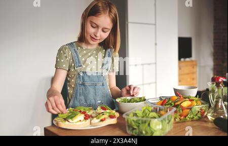 Fille préparant des sandwichs avec avocat, tomates cerises et roquette à la cuisine. Bruschettas végétariennes et salade avec légumes frais et légumes verts Banque D'Images