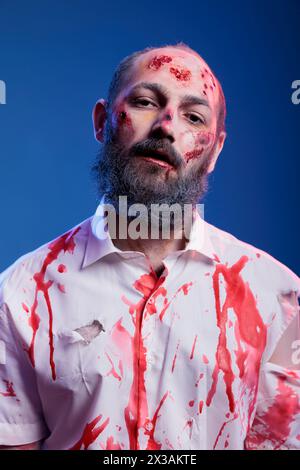 Portrait de l'acteur dépeignant le personnage de zombie dans le film d'horreur avec du sang sur le visage et les vêtements. Homme habillé en créature morte-vivante pour le film de thriller à venir, portant le maquillage de fausses blessures, fond de studio Banque D'Images