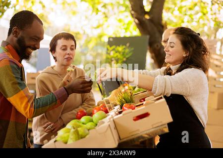 Heureux couple multiculturel dégustant des produits bio biologiques au marché des agriculteurs, achetant des fruits et légumes cultivés localement. Homme noir joyeux recevant une tranche de pomme de la vendeuse amicale. Banque D'Images