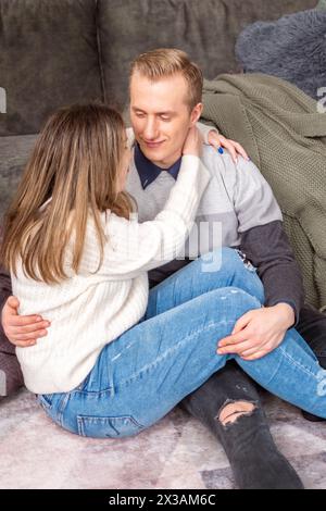 Un jeune couple partage un moment confortable sur le sol, avec les bras de l'homme tendrement enroulés autour de la femme, reflétant la chaleur et l'inclusivité de l'amour Banque D'Images