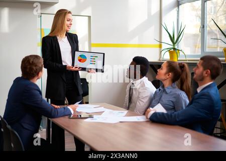 Patron féminin confiant présente des données numériques sur tablette à une équipe multiraciale attentive dans un bureau ensoleillé. Femme d'affaires dirige la réunion, discute des graphiques Banque D'Images