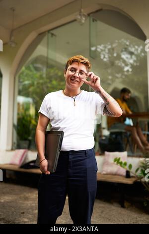 Professionnel transgenre se tient en toute confiance dans un bureau avec ordinateur portable. La pose souriante et ajustée aux lunettes reflète un environnement de travail inclusif et moderne Banque D'Images