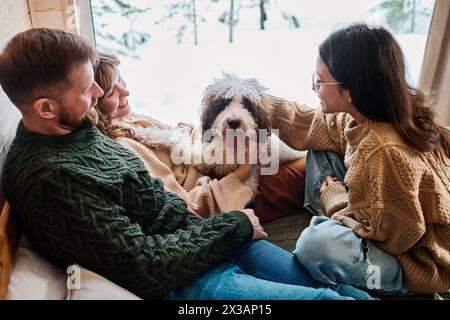 Vue en angle élevé de la famille moderne portant des chandails se relaxant avec leur chien dans un chalet en bois le jour d'hiver Banque D'Images