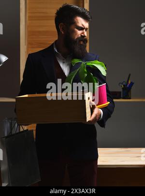 Perte d'emploi. Un employé de bureau contrarié est licencié. Chômage. Employé licencié quittant le bureau avec une boîte de ses effets personnels. Triste homme barbu en costume avec boîte Banque D'Images