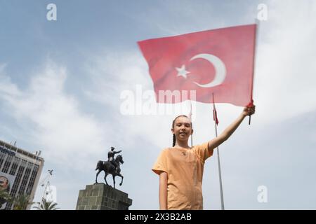 Izmir, Turquie - 23 avril 2024 : une jeune fille joyeuse agite le drapeau turc pendant les festivités de la Journée des enfants, avec la statue d'Ataturk à cheval Banque D'Images